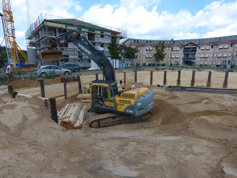 Kettenbagger in einer Baugrube mit Holzverbau neben einem Wohngebäude
